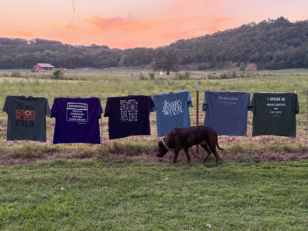T-shirts with various designs hanging on a line in a field with a dog walking by.